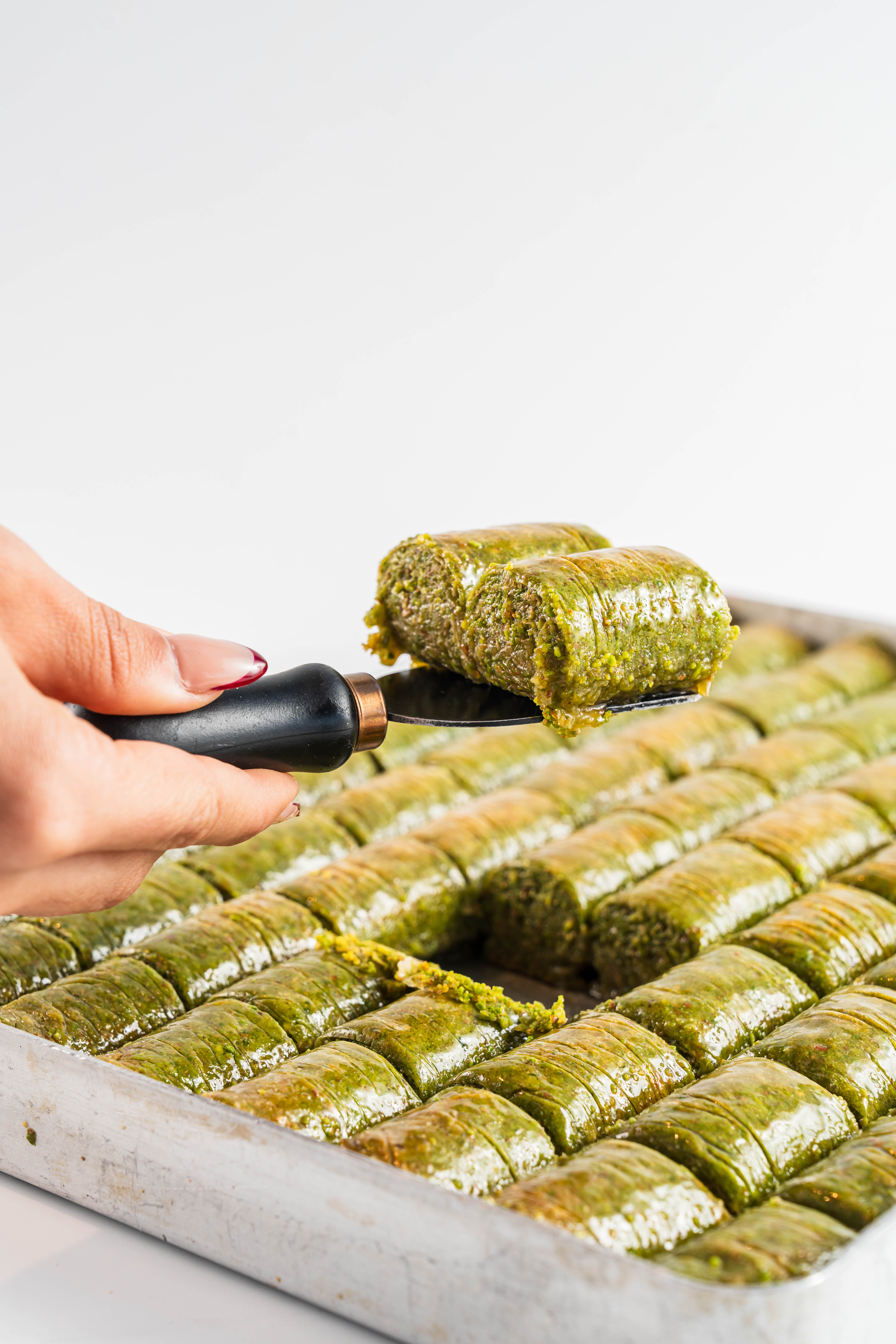 A tray of green pistachio baklava with a hand lifting two pieces on a spatula.